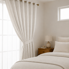 A pair of pearl white textured grommet curtains hanging on a rod in a bright, neutral-toned bedroom with a bed, nightstand, and lamp.